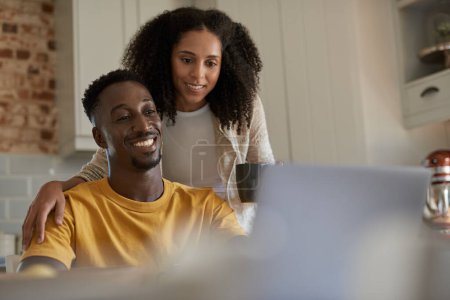 Photo for Smiling young multiethnic couple researching something on a laptop while drinking coffee together in their kitchen at home in the morning - Royalty Free Image