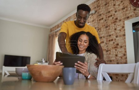 Foto de Sonriendo joven pareja multiétnica mirando algo en una tableta digital mientras beben café juntos en su cocina por la mañana - Imagen libre de derechos