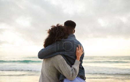 Foto de Vista trasera de una afectuosa pareja multiétnica joven de pie del brazo en una playa y mirando hacia el horizonte - Imagen libre de derechos