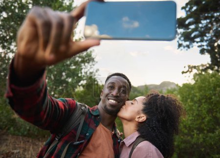 Foto de Mujer joven y cariñosa multiétnica besando a su novio sonriente en la mejilla mientras se toman una selfie juntos durante una caminata escénica - Imagen libre de derechos