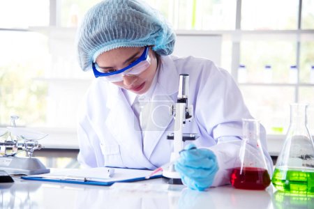 Foto de Asian Woman scientist hand write note research in medical lab look at science microscope medical test. Researcher biology chemistry. Asian Female technician laboratory using microscope blood test - Imagen libre de derechos