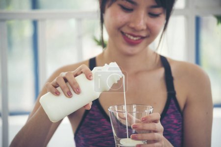 Foto de Manos de las mujeres sosteniendo vaso blanco de leche que vierte de la botella. Las mujeres asiáticas sonríen ver la cámara de atención médica estilo de vida fitness en casa. Hermosa hembra bebiendo leche fresca sin lactosa láctea baja en grasa - Imagen libre de derechos