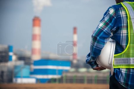 Photo for Backside Electrician engineer man hand holding White hardhat at Power stations manufacturing electrical plant. Technician worker blue hardhat helmet Engineer with steam smokestack industry background - Royalty Free Image