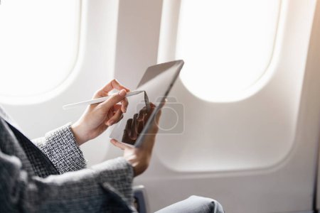 Photo for A successful asian businesswoman or female entrepreneur in formal suit in a plane sits in a business classs seat and uses a tablet computer during flight. - Royalty Free Image