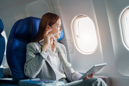 Porträt einer erfolgreichen asiatischen Geschäftsfrau oder Unternehmerin im formellen Anzug im Flugzeug sitzt auf einem Business-Class-Sitz und benutzt während des Fluges einen Tablet-Computer.