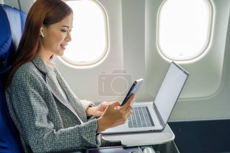 Eine erfolgreiche asiatische Geschäftsfrau oder Unternehmerin im formellen Anzug sitzt im Flugzeug auf einem Business-Class-Sitz und benutzt während des Fluges ein Smartphone.