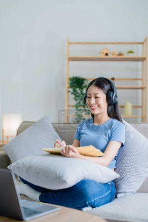 Foto de Mujer asiática joven escuchando música de auriculares y escribiendo un libro de notas para su idea de trabajo en el diario. relajar conceptos - Imagen libre de derechos