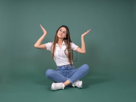 Don't know, full body front view attractive smiling woman face expression don't know. Sit floor legs crossed shrugging with hands sideways open palms. Apologizing, saying sorry. Green background. 