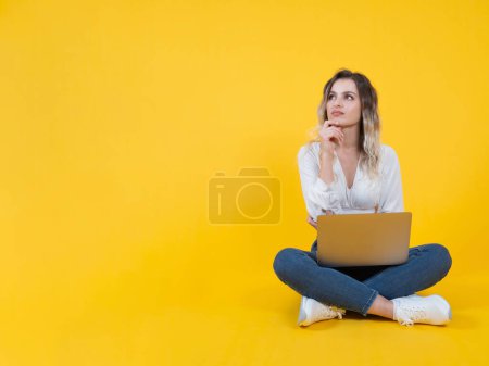 Pensive Frau, Ganzkörper junge blonde nachdenkliche Frau. Sitzen Boden isoliert gelben Hintergrund halten Laptop. Online arbeiten, freiberuflich. Den Blick zur Seite gelegt und das Kinn aufgestützt. Lebensstil-Konzept der Menschen.