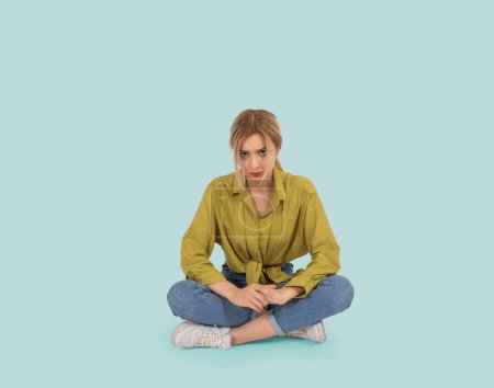 Offended angry young woman, full body length portrait of offended angry young woman. Sit floor legs crossed. Upset, unhappy conflict looking blonde girl. Isolated light blue background. Copy space.