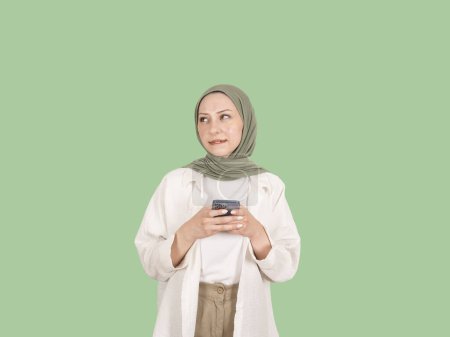 Téléchargez les photos : Femme musulmane rêveuse, portrait de jeune femme musulmane songeuse caucasienne portant des vêtements causals et du hijab. Tenant le téléphone, posant sur fond vert. Regarder l'espace de copie, mordre les lèvres. Mode de vie. - en image libre de droit