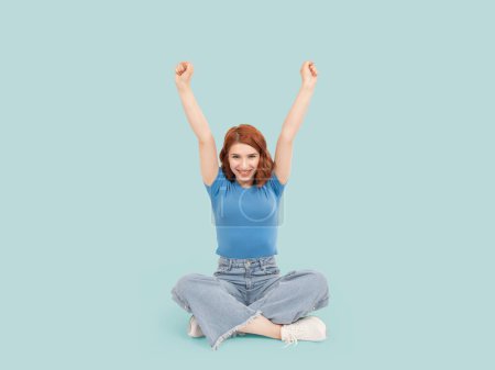 Woman celebrating, full body size funny happy caucasian red bob hair woman celebrating. Wear jeans and blue t shirt. Sit floor isolated blue studio background. Clenching fists, win bet concept idea.