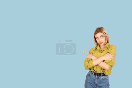 Serious confident young 20s caucasian blonde woman pointing aside, stand over blue background copy space. Crossing arms, negative emotion expression angry close body language.