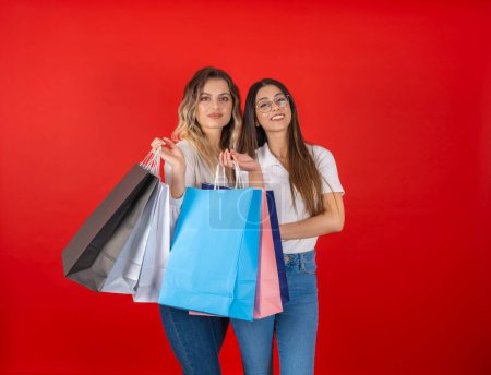 Porträt zweier glücklicher junger Frauen mit Einkaufstüten, die nach einem langen, angenehmen Tag im Einkaufszentrum vor rotem Studiohintergrund posieren. Kaukasische schöne modische Freundinnen lächelnde Kamera. 