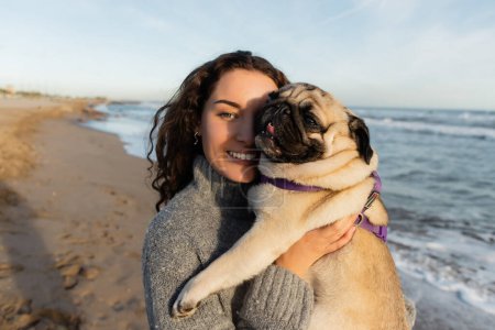 cheerful and curly young woman holding pug dog on beach near sea in Barcelona 