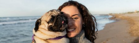 fröhliche und lockige junge Frau mit Mops-Hund am Strand am Meer, Banner 