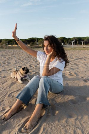 lockige junge Frau sitzt mit ausgestreckter Hand neben Mops-Hund am Sandstrand in Barcelona 
