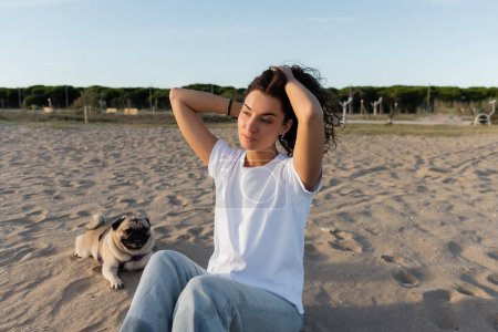 young woman adjusting curly hair while sitting near pug dog on sandy beach in Barcelona 