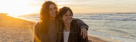 Téléchargez les photos : Femme gaie étreignant ami et souriant tout en marchant pendant le coucher du soleil sur la plage de sable, bannière - en image libre de droit
