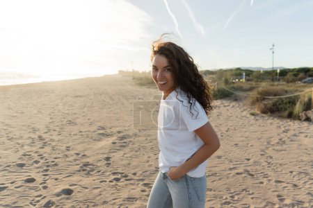 alegre joven en camiseta blanca caminando por la playa de arena en España 