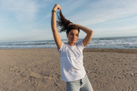 femme brune en t-shirt blanc ajustant les cheveux bouclés sur la plage de sable près de la mer 