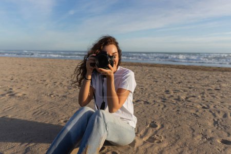 lockige Frau sitzt in blauen Jeans und weißem T-Shirt und fotografiert mit Digitalkamera in Spanien 