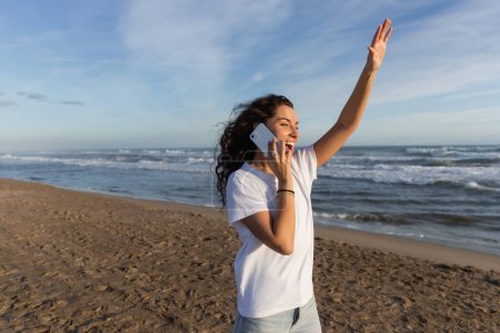 excited woman in white t-shirt talking on smartphone and waving hand on beach in Barcelona 