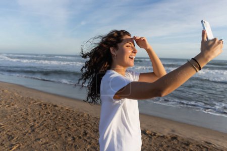 curly woman in white t-shirt taking selfie on smartphone on sandy beach in Barcelona 