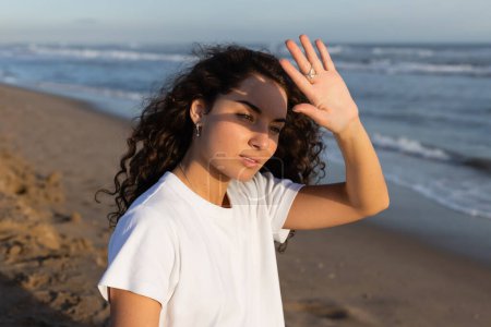 Porträt einer lockigen Frau im weißen T-Shirt, die ihr Gesicht vor der Sonne am Strand am Meer versteckt 