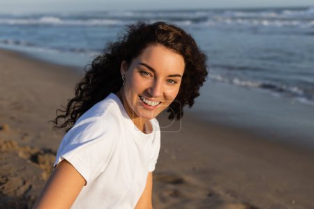Téléchargez les photos : Portrait de femme positive et bouclée en t-shirt blanc sur une plage de sable près de la mer - en image libre de droit