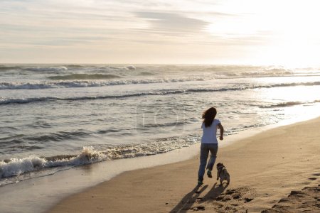 Photo pour Vue arrière de la femme courant avec chiot chiot sur la plage de sable fin pendant le coucher du soleil - image libre de droit