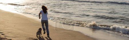 Téléchargez les photos : Vue arrière de la femme courant avec chiot chiot sur la plage de sable fin pendant le coucher du soleil, bannière - en image libre de droit