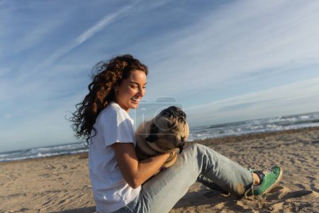happy young woman with curly hair holding pug dog while sitting on beach near sea in Spain 