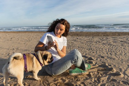 cheerful woman in white t-shirt taking photo of pug dog on sandy beach in Barcelona 