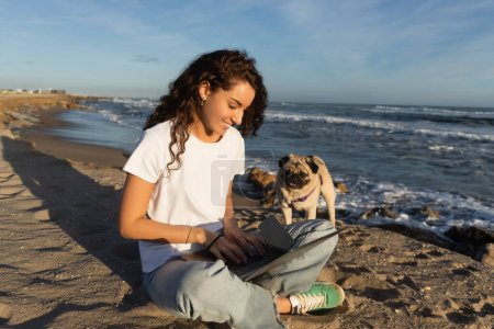 glückliche junge Freiberuflerin mit lockigem Haar mit Laptop in der Nähe von Mops Hund am Strand am Meer in Spanien 
