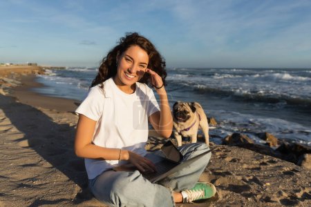 fröhliche junge Freiberuflerin mit lockigem Haar mit Laptop in der Nähe von Mops Hund am Strand am Meer in Spanien 
