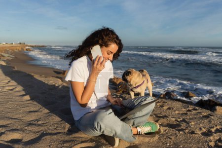 cheerful freelancer talking on smartphone while using laptop near pug dog on beach near sea in Spain 