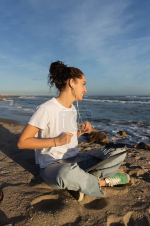 jeune freelance souriant dans des écouteurs filaires assis avec ordinateur portable sur la plage à Barcelone