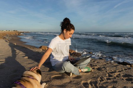 Téléchargez les photos : Young freelancer in wired earphones sitting with laptop and cuddling pug dog on beach in Barcelona - en image libre de droit