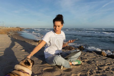 young woman in wired earphones holding smartphone near laptop and cuddling pug dog on beach in Barcelona magic mug #634412418