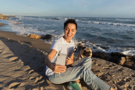 Téléchargez les photos : Happy woman in wired earphones holding laptop and taking selfie with pug dog on beach in Barcelona - en image libre de droit