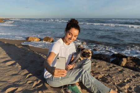 Téléchargez les photos : Cheerful woman in wired earphones holding laptop and taking selfie with pug dog near sea in Spain - en image libre de droit
