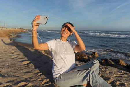 Téléchargez les photos : Cheerful woman in wired earphones sitting with laptop and taking selfie near sea in Spain - en image libre de droit