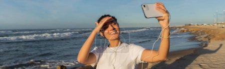 fröhliche Frau mit kabelgebundenen Kopfhörern macht Selfie am Meer in Spanien, Banner