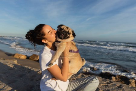 young woman with curly hair kissing pug dog on beach near sea in Barcelona 