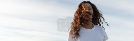 young woman with curly hair looking at camera against sky in Barcelona, banner 