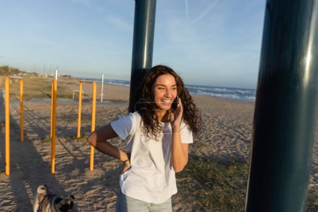 fröhliche junge Frau spricht mit Smartphone, während sie mit der Hand auf der Hüfte am Strand in Barcelona steht 
