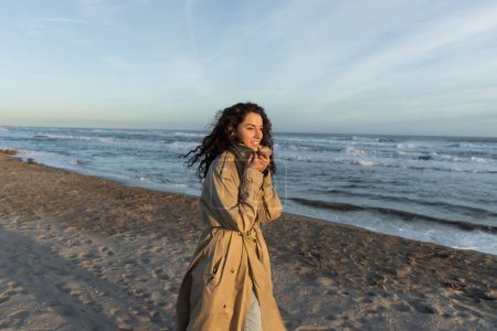 cheerful woman in stylish beige trench coat walking on beach in Barcelona 