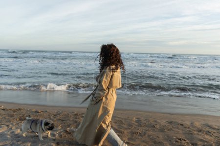 full length of curly woman in trench coat walking with pug dog on beach near sea in Barcelona 