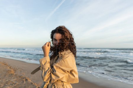 young woman in beige trench coat adjusting curly hair while looking at camera near sea in Barcelona 